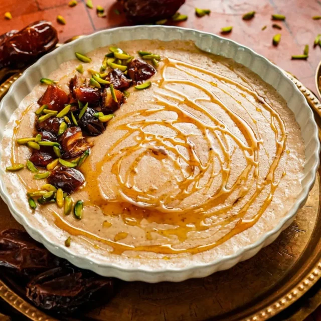 A bowl of creamy Talbina topped with chopped dates, slivered pistachios, and a drizzle of honey, placed on a brass tray with extra dates around.